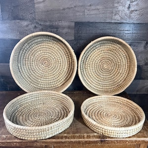 May include: Four round, woven baskets in varying sizes. The baskets are made of a light tan material and have a spiral design in the center. The baskets are displayed on a wooden surface against a dark wood background.