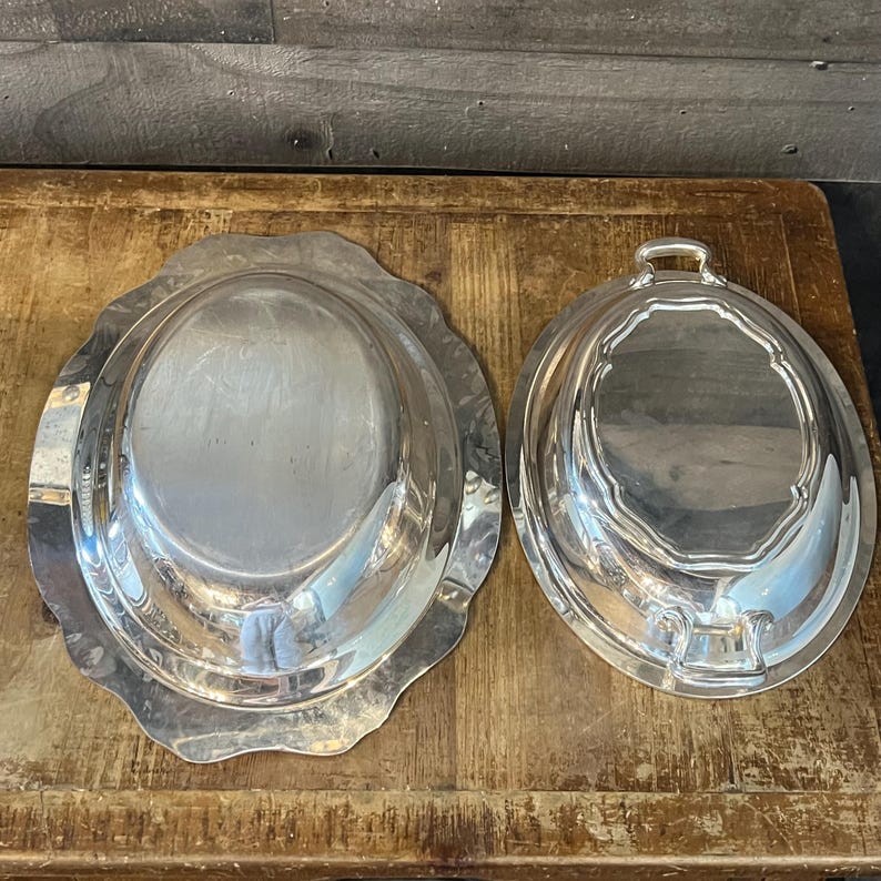 May include: Two silver serving dishes with lids. One dish has a scalloped edge and an oval shape. The other dish has a handle and a matching lid. Both dishes are on a wooden surface.