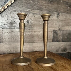 May include: A pair of gold metal candle holders with a simple, elegant design. The holders have a tall, slender base and a wide, round top.