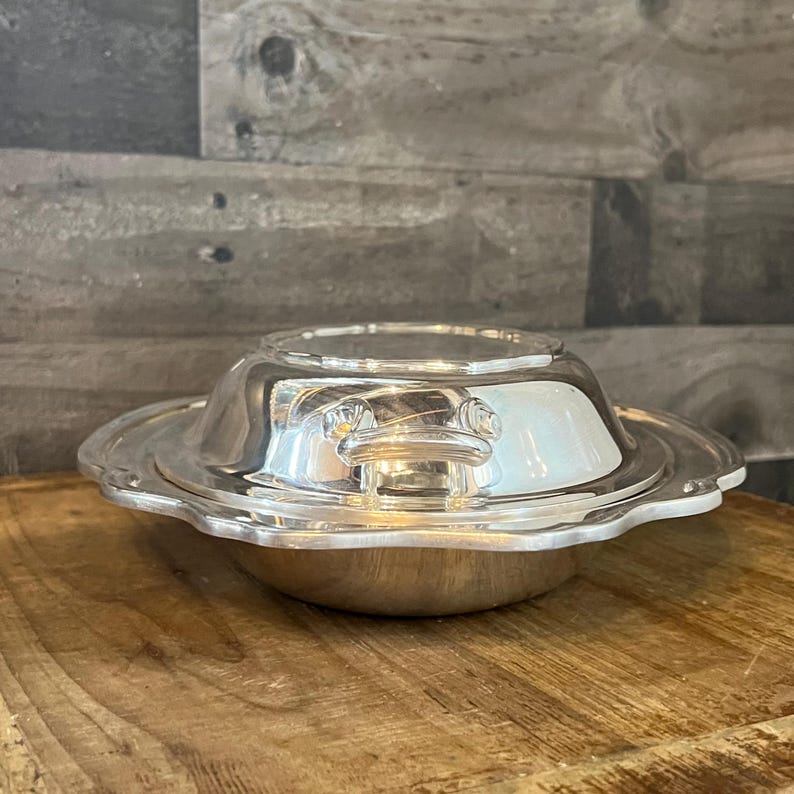 May include: A silver-colored, covered serving dish with a scalloped edge. The dish has a handle on the lid and a rounded base. The dish is sitting on a wooden surface.
