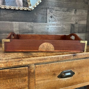 Puede incluir: Una bandeja rectangular de madera con un acabado marrón rojizo oscuro. La bandeja tiene esquinas de latón y una placa de latón con el texto "TANIC 1913". Cuenta con asas recortadas y se encuentra sobre una superficie de madera.