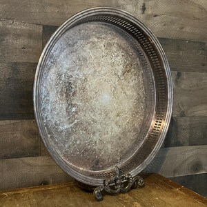 May include: Oval silver serving tray with a decorative pierced border and ornate detailing. The tray has a tarnished appearance, suggesting age and use. A decorative metal stand with a fleur-de-lis design supports the tray.