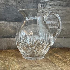 May include: A clear glass pitcher with a handle. The pitcher has a diamond-cut pattern on the lower portion and a smooth, curved design on the upper portion. The pitcher is sitting on a wooden surface.