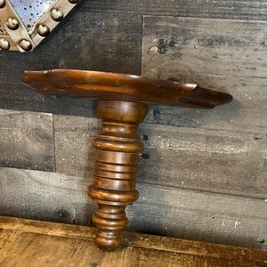 May include: A dark brown, antique-style wooden shelf with a decorative, turned pedestal base. The shelf has a curved, organic shape and is mounted on a wall. The wood grain is visible, adding to the vintage aesthetic. The shelf is approximately 24 inches long.