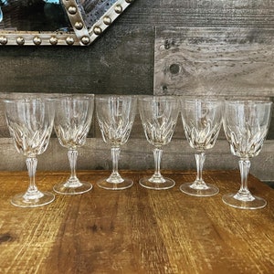May include: Six clear crystal wine glasses with a decorative etched pattern. The glasses have a classic design with a stemmed base and a wide bowl. The glasses are arranged on a wooden surface.