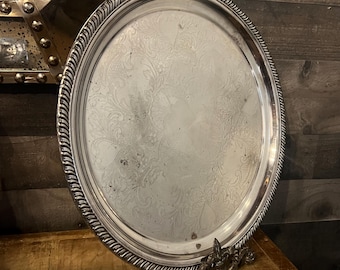 May include: Silver-plated oval serving tray with a decorative rope-like edge and ornate floral pattern. The tray has a decorative base and is displayed on a wooden surface. A vintage piece for serving or display.