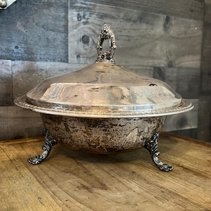 May include: A silver serving dish with a lid and three ornate feet. The dish has a tarnished finish and is sitting on a wooden surface.