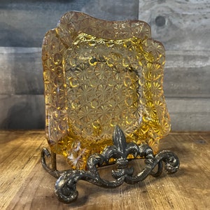 Könnte beinhalten: Ein antiker Aschenbecher aus Glas mit einem dekorativen Blumenmuster steht auf einem Metallständer. Der Aschenbecher ist klar und hat eine gelbe Tönung. Der Ständer ist verziert und hat ein Fleur-de-lis-Design.