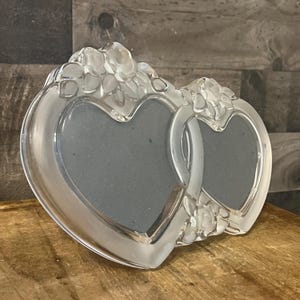 May include: A crystal picture frame with two interlocking heart-shaped openings. The frame is decorated with floral details.