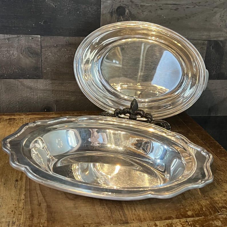 May include: Two silver-colored serving dishes. The bottom dish has a scalloped edge. The top dish is oval-shaped. A black decorative metal stand is between the two dishes. The dishes are on a wooden surface.