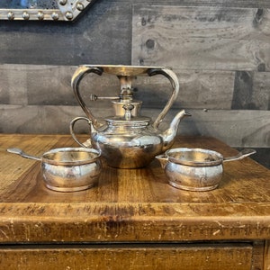 Può includere: Una teiera d'argento con un supporto d'argento e due brocche per la panna d'argento su un tavolo di legno.