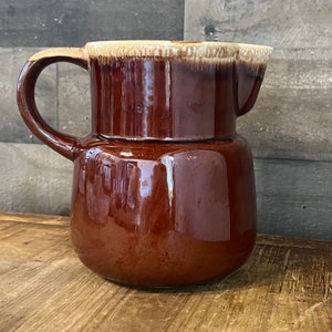 Puede incluir: Una jarra de cerámica marrón con un borde marrón claro y un asa. La jarra tiene un acabado brillante.
