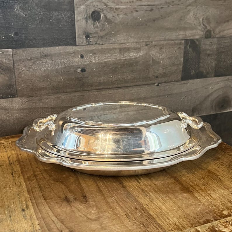 May include: A silver-colored serving dish with a lid. The dish has a scalloped edge and ornate handles on either side. The dish is sitting on a wooden surface.