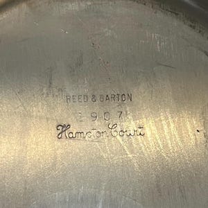 May include: Close-up of a silver-toned metal surface with the engraved text "REED & BARTON 1907 Hampton Court". The surface has a slightly textured appearance with some visible scratches, suggesting age and use. The text is in a classic serif font.