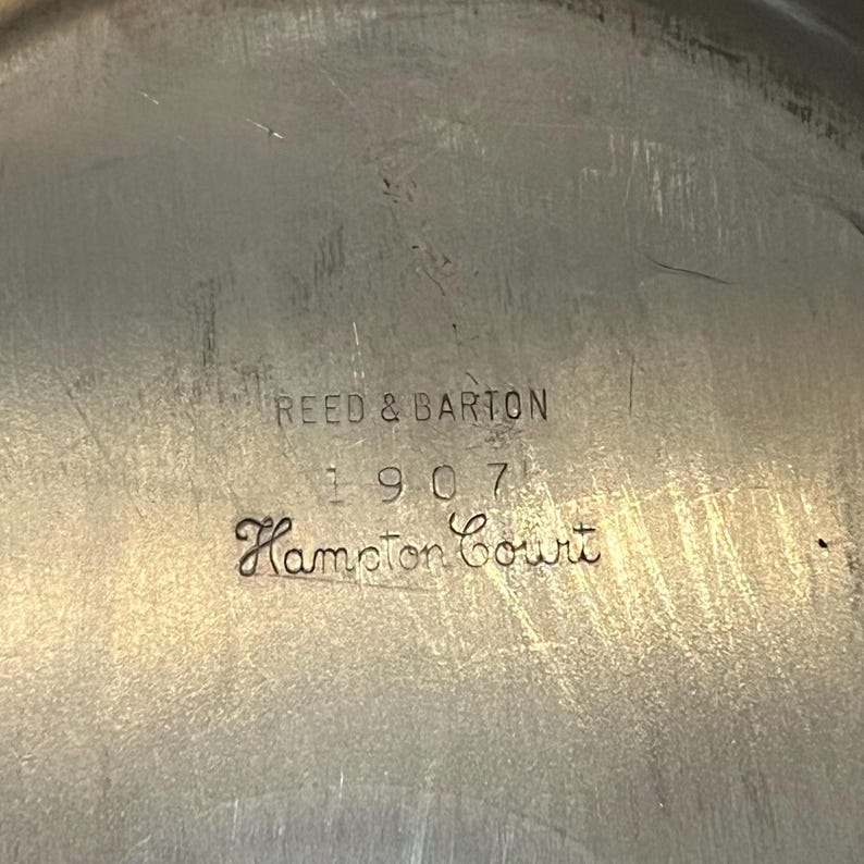 May include: Close-up of a silver-toned metal surface with the engraved text "REED & BARTON 1907 Hampton Court". The surface has a slightly textured appearance with some visible scratches, suggesting age and use. The text is in a classic serif font.