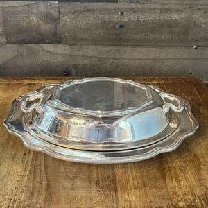 May include: A silver-colored, oval-shaped serving dish with a lid. The dish has a decorative, scalloped edge and a reflective surface. The lid fits snugly on the base, creating a complete serving piece. The dish is on a wooden surface.