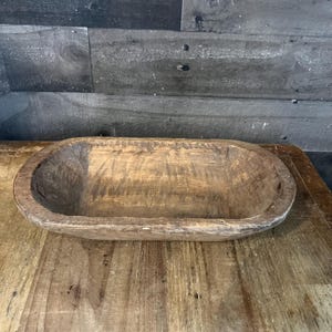 May include: A rustic wooden dough bowl with a natural wood finish. The bowl is oval shaped and has a smooth, slightly textured surface.