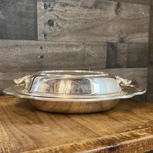 May include: A silver-colored, covered serving dish with ornate handles and a scalloped edge. The dish is round and reflective, sitting on a wooden surface. The lid is slightly domed, and the handles are curved.