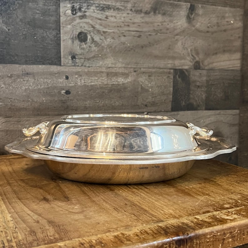 May include: A silver-colored, covered serving dish with ornate handles and a scalloped edge. The dish is round and reflective, sitting on a wooden surface. The lid is slightly domed, and the handles are curved.