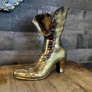May include: A gold-toned metal boot sculpture with a high heel and a buttoned flap. The boot is made of a shiny metal and has a vintage style.