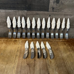 May include: A set of 14 white seashell-shaped forks and butter knives. The forks and knives are arranged in two rows, with the forks at the front and the knives at the back. The forks and knives are all made of metal and have a polished finish.