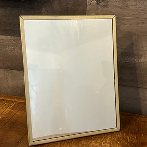 Puede incluir: Un marco de fotos dorado con un paspartú blanco y un interior blanco vacío. El marco está sobre una superficie de madera.