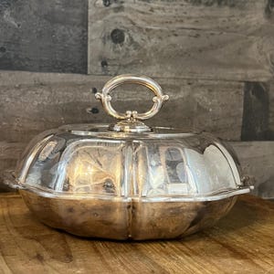 Puede incluir: Una fuente de servir de plata con tapa abovedada y asa decorativa. La fuente tiene un borde festoneado y un acabado brillante.