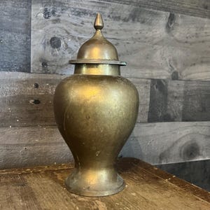 May include: A vintage brass urn with a lid. The urn has a bulbous body and a conical top, with a small finial. The brass has a warm, aged patina. The urn is displayed on a wooden surface against a wood-paneled wall.