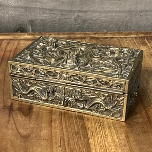 Puede incluir: Una caja de latón con intrincados diseños de dragones y flores. La caja está cerrada y tiene una tapa con bisagras.