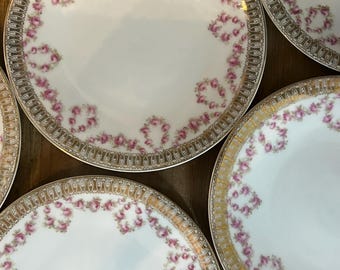 May include: Several white porcelain plates with gold rims and pink rose garlands. The plates are displayed on a wooden surface, highlighting their floral design and vintage style. Each plate has a diameter of approximately 20 cm.