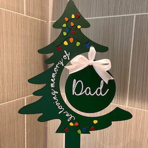 May include: A green Christmas tree cutout with colourful lights and a white ribbon ornament that says "Dad" in white lettering. The tree is on a stick and the text "In loving memory of" is written on the tree.