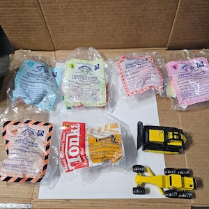 May include: Five Happy Meal toy wrappers from McDonald's featuring a yellow Tonka Loader toy, a yellow Tonka Loader toy, a pink and white Happy Meal wrapper with the text "Congratulations on your new arrival!" and a blue and white Happy Meal wrapper with the text "Congratulations on your new arrival!" and a yellow and white Happy Meal wrapper with the text "Congratulations on your new arrival!"
