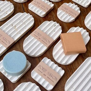 May include: A collection of white ceramic soap dishes with a textured, speckled finish. The dishes are oval and rectangular in shape and have raised ridges on the bottom. Some of the dishes have the text "PORTE-SAVON" printed on them.