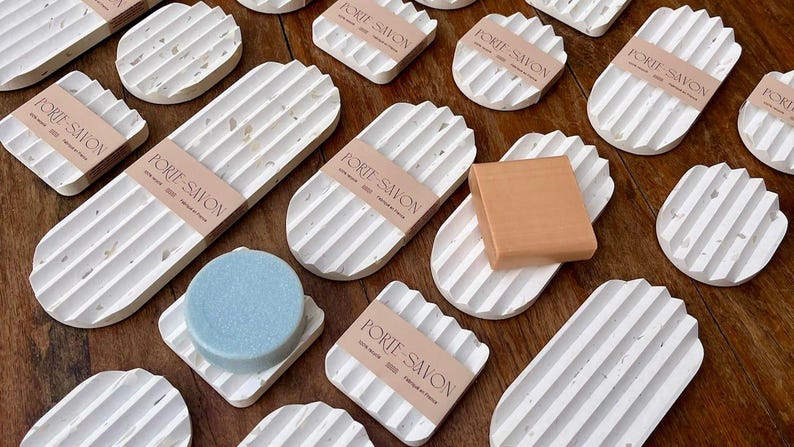 May include: A collection of white ceramic soap dishes with a textured, speckled finish. The dishes are oval and rectangular in shape and have raised ridges on the bottom. Some of the dishes have the text "PORTE-SAVON" printed on them.