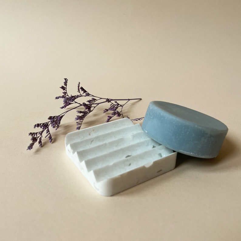 May include: A white soap dish with a textured surface and a grey soap bar on top. The soap dish is made of ceramic and has a rectangular shape. A sprig of dried purple flowers is in the background.
