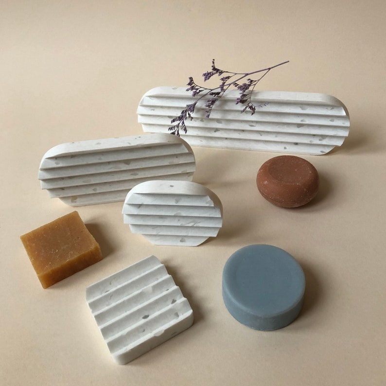 May include: Five white soap dishes with a speckled texture and a grooved design. Two of the dishes are oval shaped, one is round, and two are rectangular. There is a brown soap bar and a blue soap bar on the dishes.