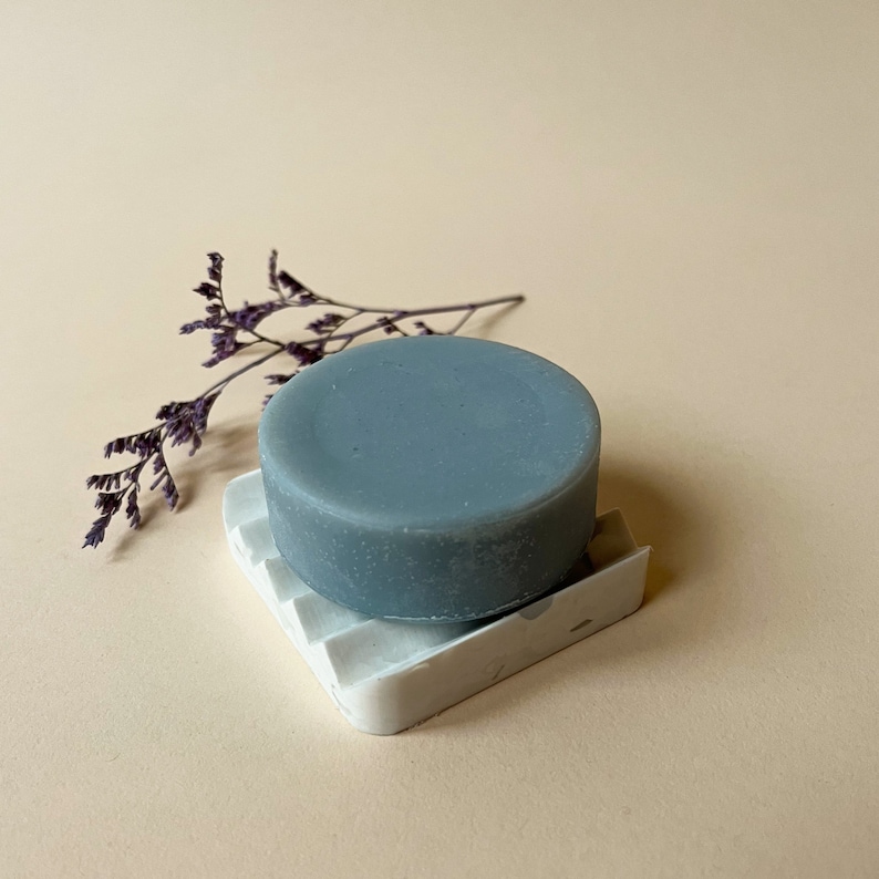 May include: A round, grey soap bar sits on a white, textured soap dish. The soap dish has a raised, textured design. A sprig of dried purple flowers is in the background.