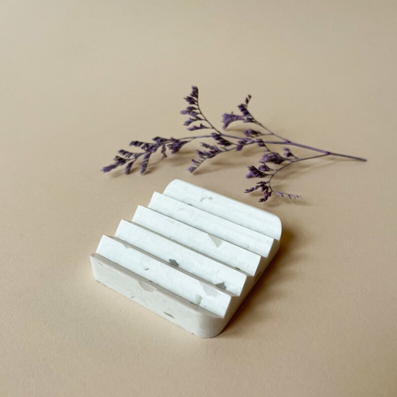 May include: White ceramic soap dish with a textured surface and a rippled design. The dish is made of speckled concrete.