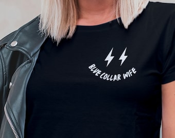 Może przedstawiać: Czarny t-shirt z białym napisem "BLUE COLLAR WIFE" nad dwoma białymi błyskawicami. Czarna skórzana kurtka jest zarzucona na ramiona.