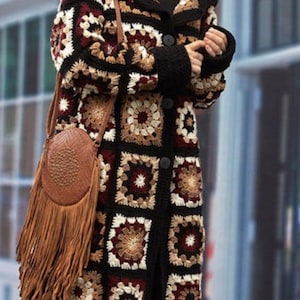 May include: A long, handmade crochet coat in a patchwork design. The coat features a black base with squares of cream, brown, and burgundy. A brown leather shoulder bag with fringe hangs on the side. The coat has a button closure and a hood.