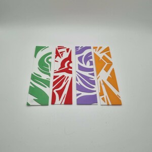 Peut inclure: Quatre marque-pages rectangulaires disposés côte à côte. Chaque marque-page présente un motif découpé unique dans une couleur unie : vert, rouge, violet et orange. Les découpes créent des motifs abstraits sur la base blanche de chaque objet.