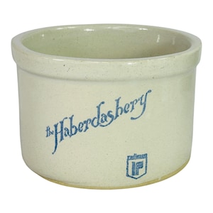 May include: A cream-colored stoneware crock with a wide opening. The blue text on the side reads "The Haberdashery" with a logo below. The crock has a slightly speckled appearance and a rounded base.