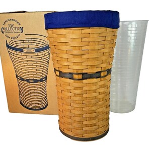 May include: A Longaberger J.W. Collection 1994 Edition umbrella basket. The basket is made of woven light brown wood with a dark blue fabric top and a black band. A clear plastic container is next to the basket. The box has a drawing of the basket.