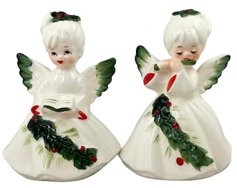 May include: Two white ceramic angel figurines with green wings and holly garland accents on their dresses. One angel holds an open book, while the other plays a flute. Both figures feature painted facial details, red lips, and holly in their hair.