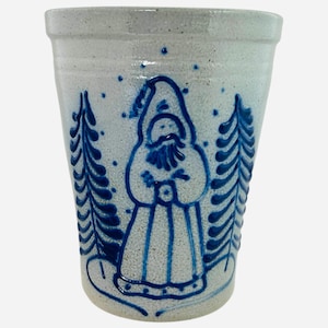 May include: A stoneware crock with a light gray background and a blue Santa Claus design. The Santa Claus is wearing a hooded robe and is flanked by two blue Christmas trees. The crock has a slightly flared shape and a textured surface.