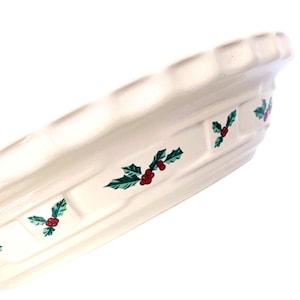 May include: Cream-colored ceramic serving dish with a scalloped edge and a woven basket design. The dish is decorated with green holly leaves and red berries, perfect for holiday gatherings.
