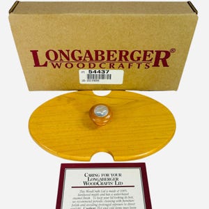 May include: A wooden Longaberger Woodcrafts lid with a light brown finish. The lid has an oval shape with a small handle and a cutout. The lid is part of a set, with a box and care instructions visible.