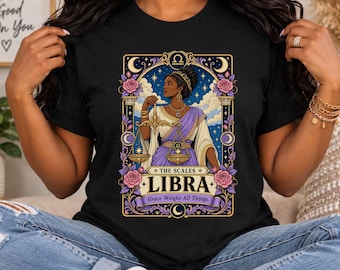 May include: A black t-shirt featuring a graphic of a person holding a golden scale, surrounded by pink roses, stars, and crescent moons. The text reads THE SCALES LIBRA Grace Weighs All Things. The design is framed by ornate pillars and celestial symbols.