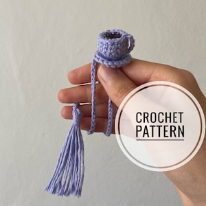 Può includere: Una tazza e un piattino all'uncinetto color lavanda con un fiocco appeso al manico. Il testo "CROCHET PATTERN" è visibile nell'immagine.