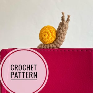 May include: Crochet pattern for a small brown and yellow snail. The snail is sitting on a pink notebook with the words "Crochet Pattern" in a white circle.